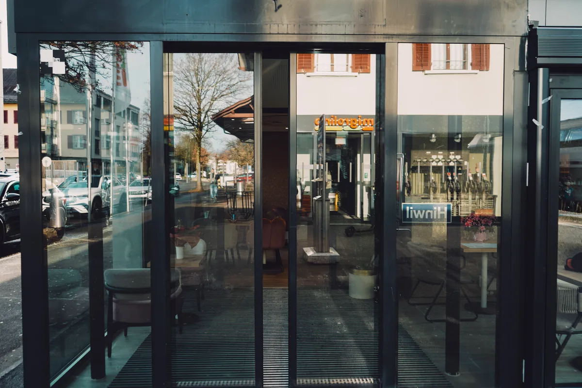 Man sieht die Eingangstür zu einem Café. In der Spiegelung ist der Schriftzug Hinwil zu lesen.